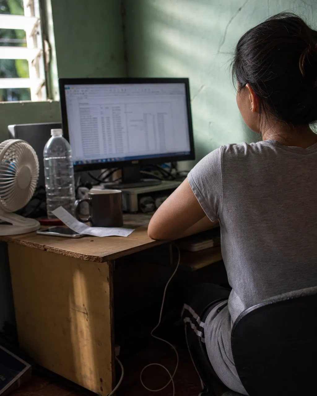Seen from behind, a woman works at a monitor at a cluttered home desk beside a jalousie window in early-morning light.