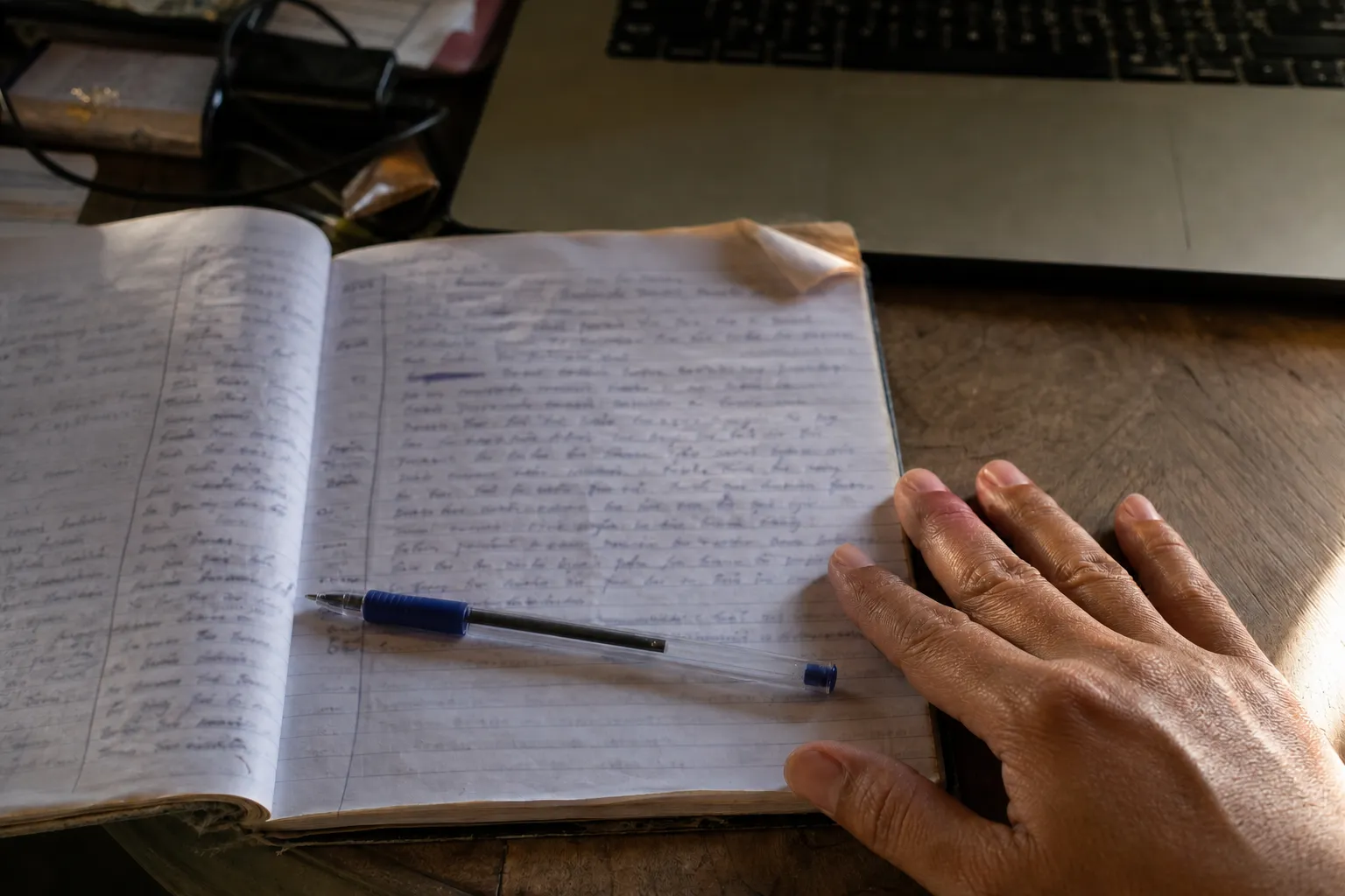 A close view of a well-used notebook with handwritten notes softened out of focus, a pen lying on the page and a hand resting at its edge.