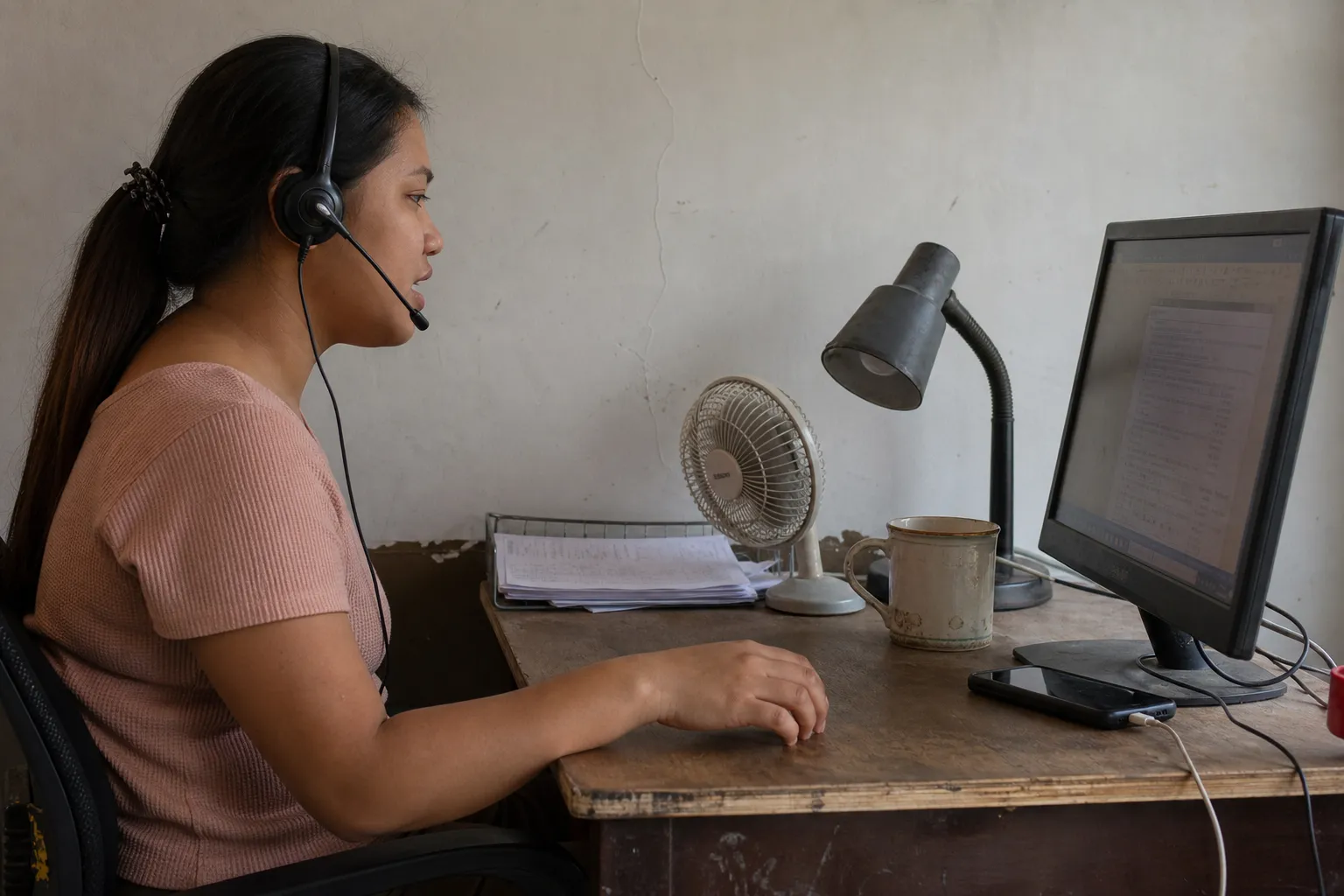 In profile, a woman on a headset speaks mid-sentence at a desk, a switched-off lamp, a tray of papers and a desk fan beside her monitor.