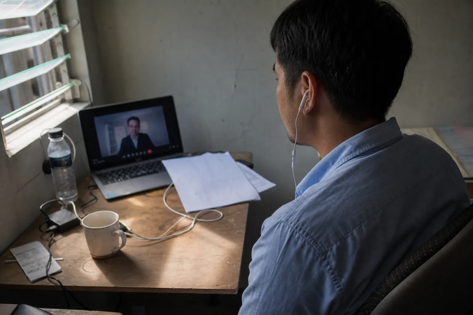 Seen from behind, a man takes a video interview on a laptop at a plywood desk, wired earphones in and a printed CV face-down beside him.