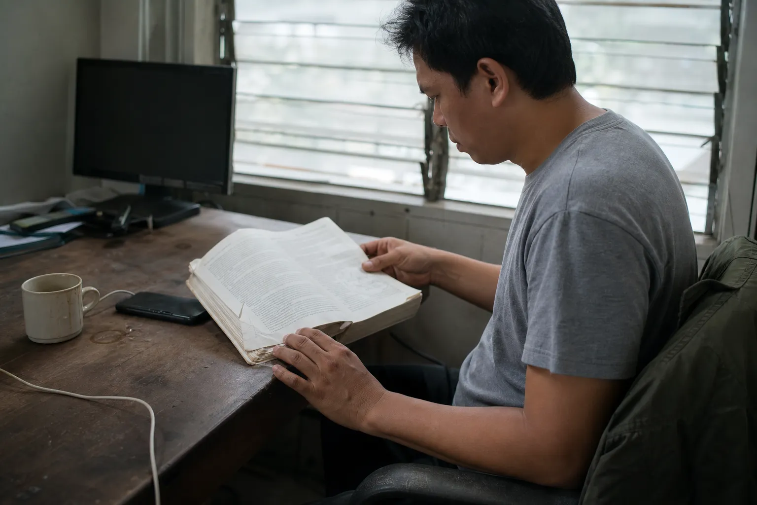 A man reads a well-used printed manual at a desk, seen from the side with his head down, an overcast window behind him.