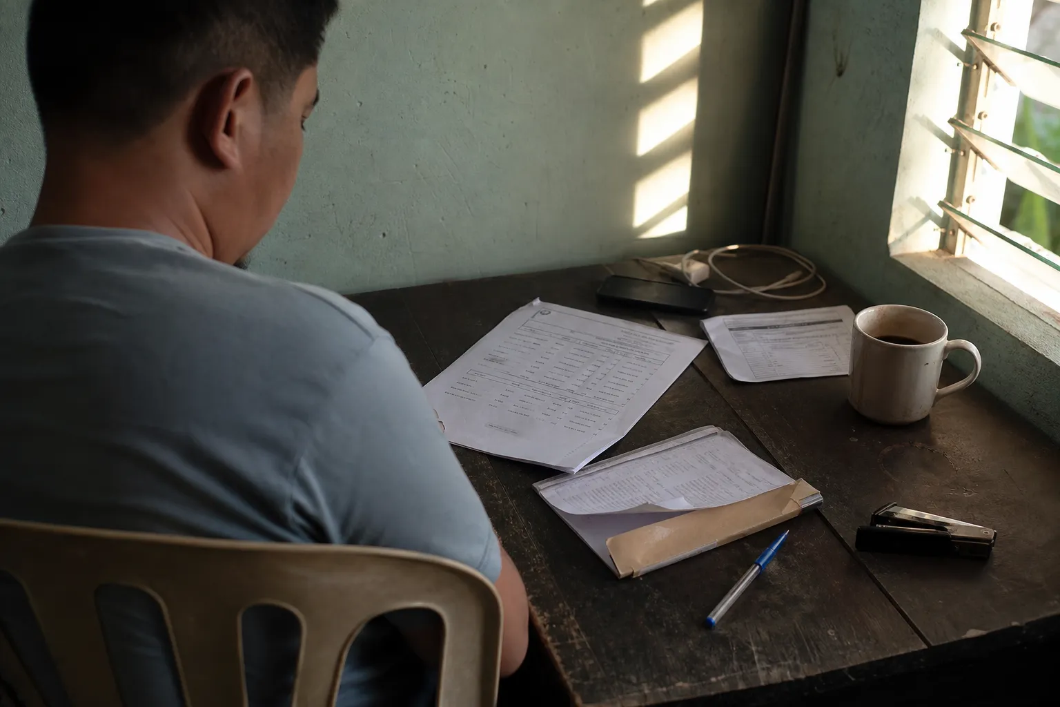 Seen from behind, a man reads a printed sheet at a small table in a home office, his phone face-down beside a mug in morning light.